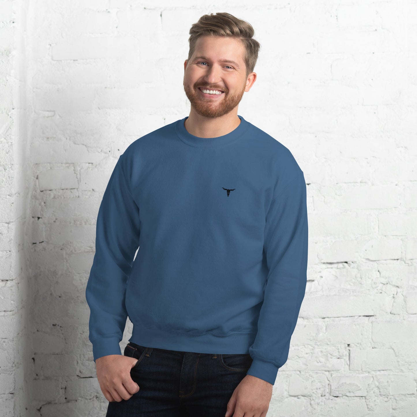 Man wearing blue Bull Skull unisex sweatshirt with classic fit and small bull skull logo on chest against white brick wall background