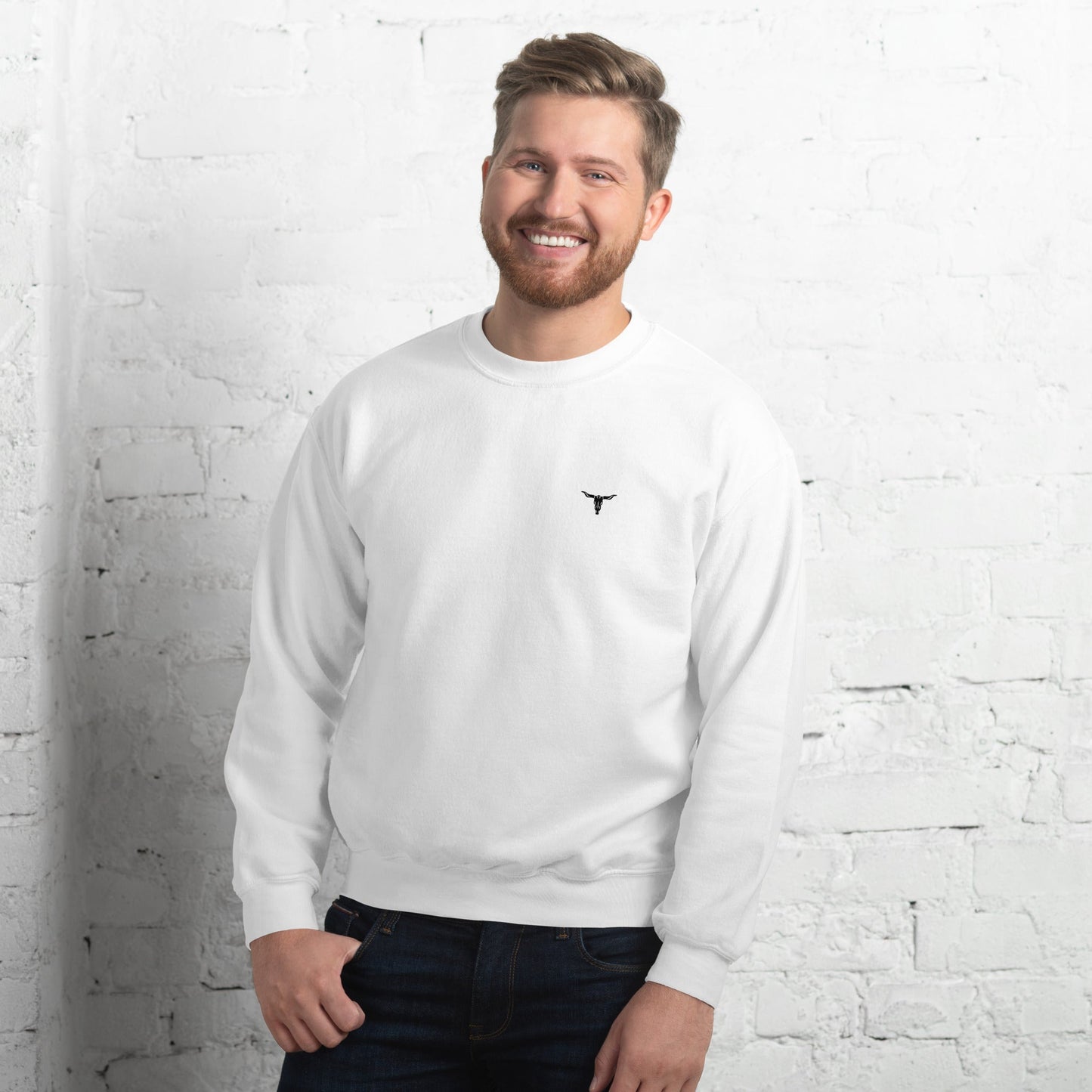 Man wearing white Bull Skull Unisex Sweatshirt with black bull skull logo on chest standing against white brick wall