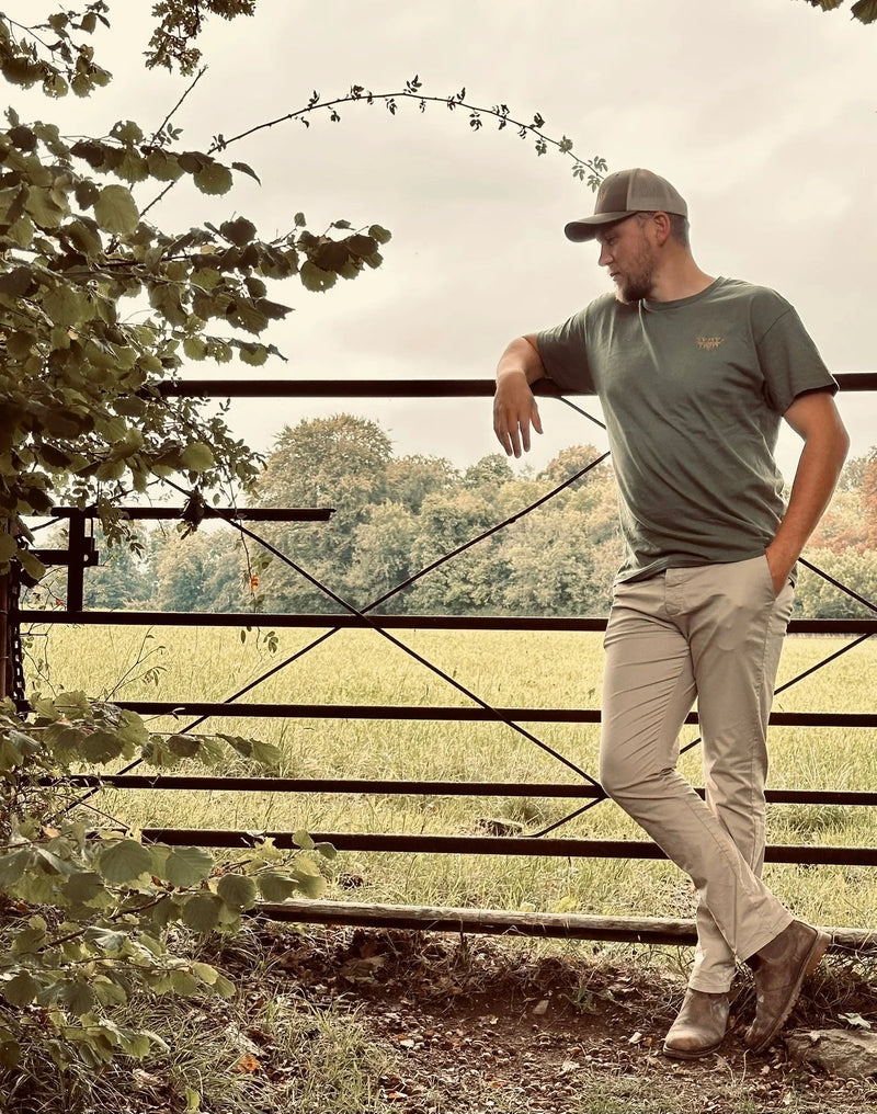 Man in green t-shirt and cap leaning on metal gate in rural field with trees in background