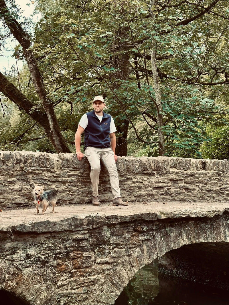 Man in casual outdoor clothing and small dog standing on rustic stone bridge in lush green forest