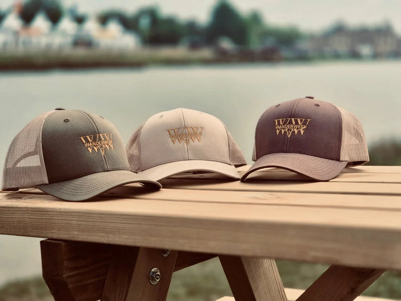 WanderWest logo trucker hats in earthy colors on a wooden picnic table by a lake