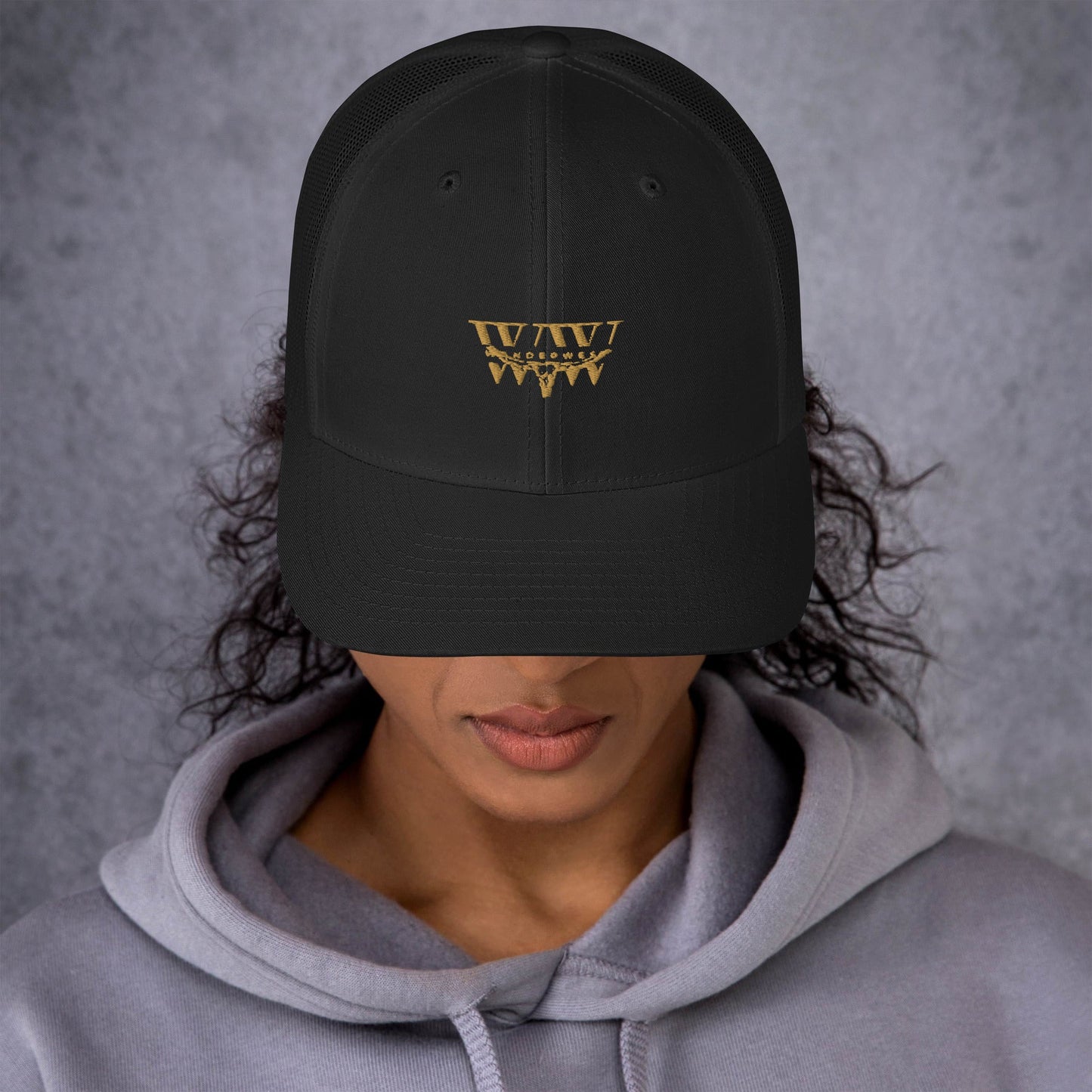 Black WanderWest Logo Trucker Cap with gold embroidery, mesh back, mid-profile fit, worn by person in gray hoodie.