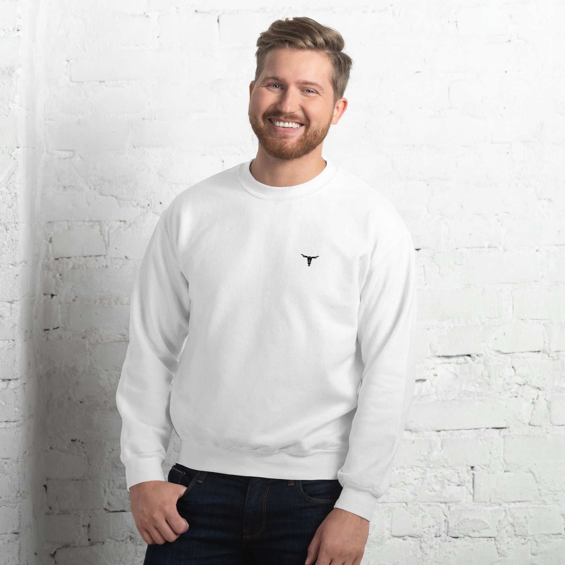 Man wearing white Bull Skull Unisex Sweatshirt with black bull skull logo on chest standing against white brick wall